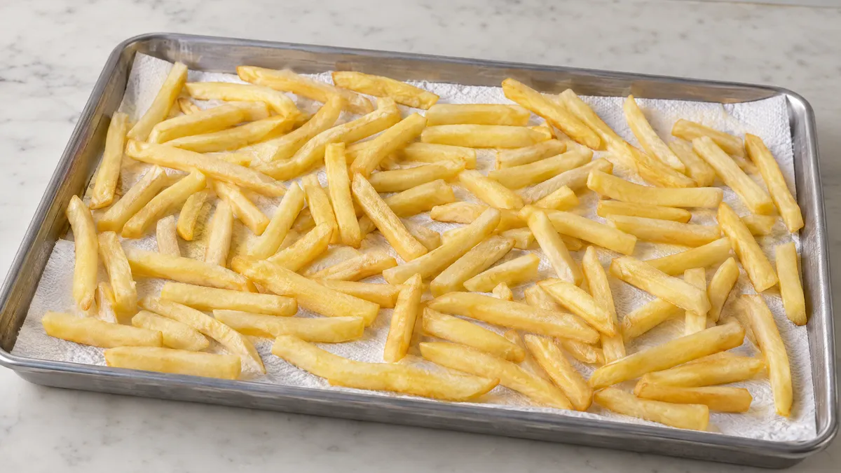 Blanched fries cooling uncovered in a single layer on a tray lined with paper towels.