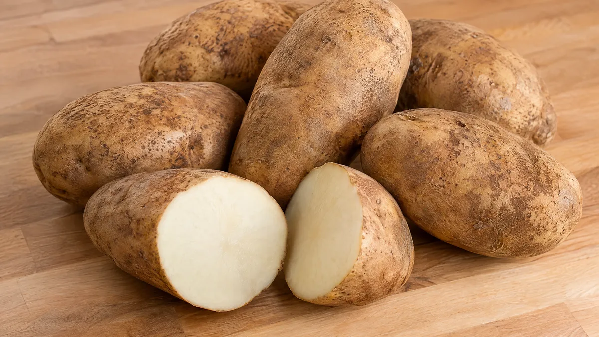 Whole russet potatoes on a wooden counter, one cut in half to show the white flesh.
