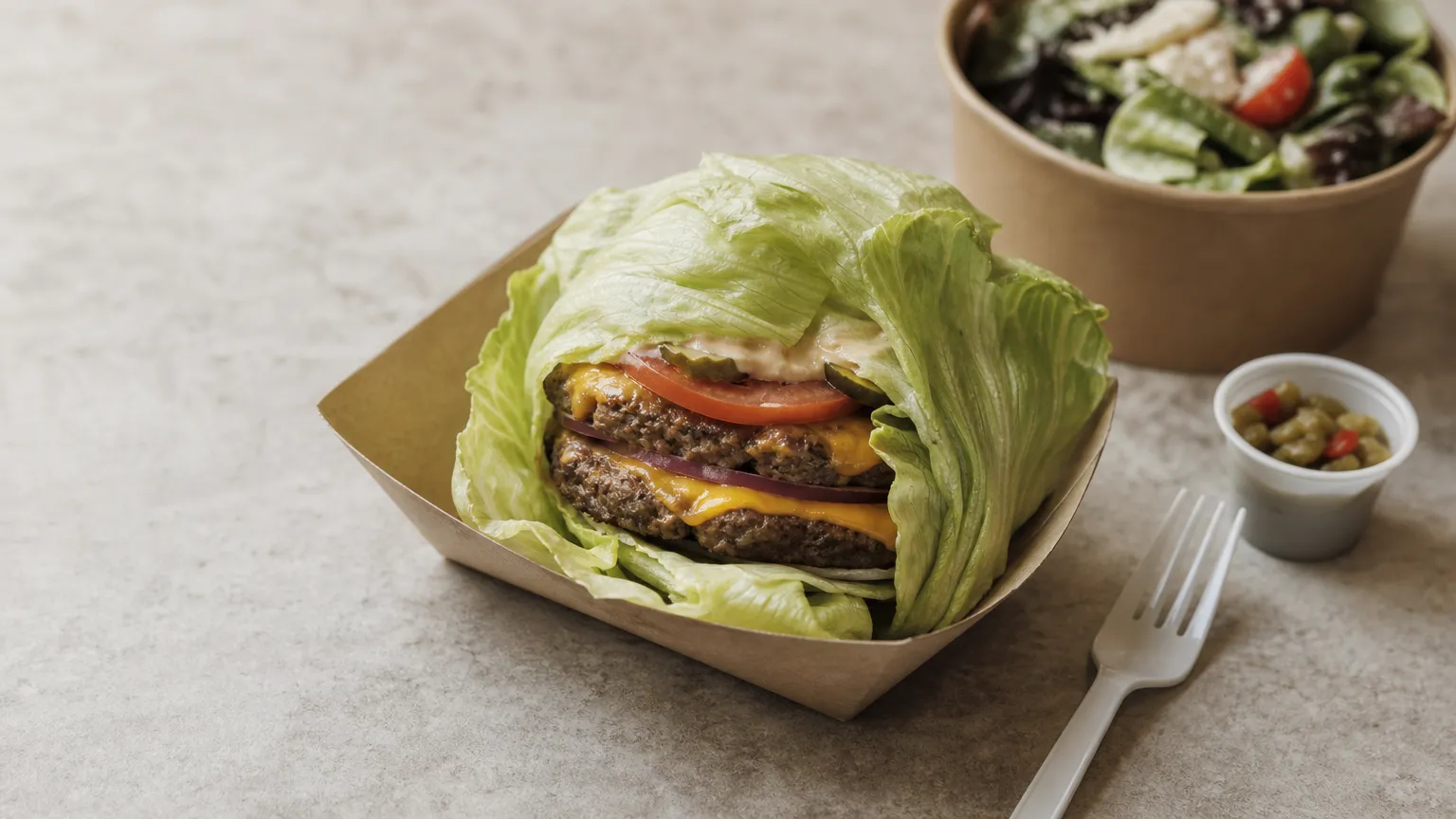 Lettuce-wrapped double burger on a paper tray with a fork, chili cup and side salad.